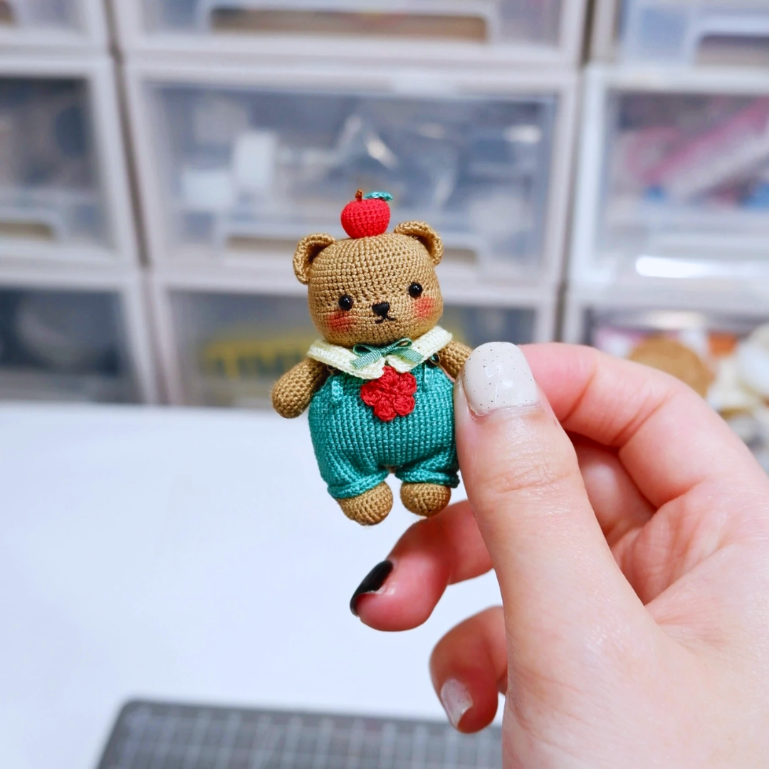 Handheld brown micro crochet bear charm with apple detail – cute handmade gift accessory.