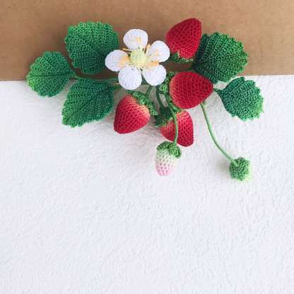 Crocheted strawberry bush with green leaves and red strawberries on a white surface.