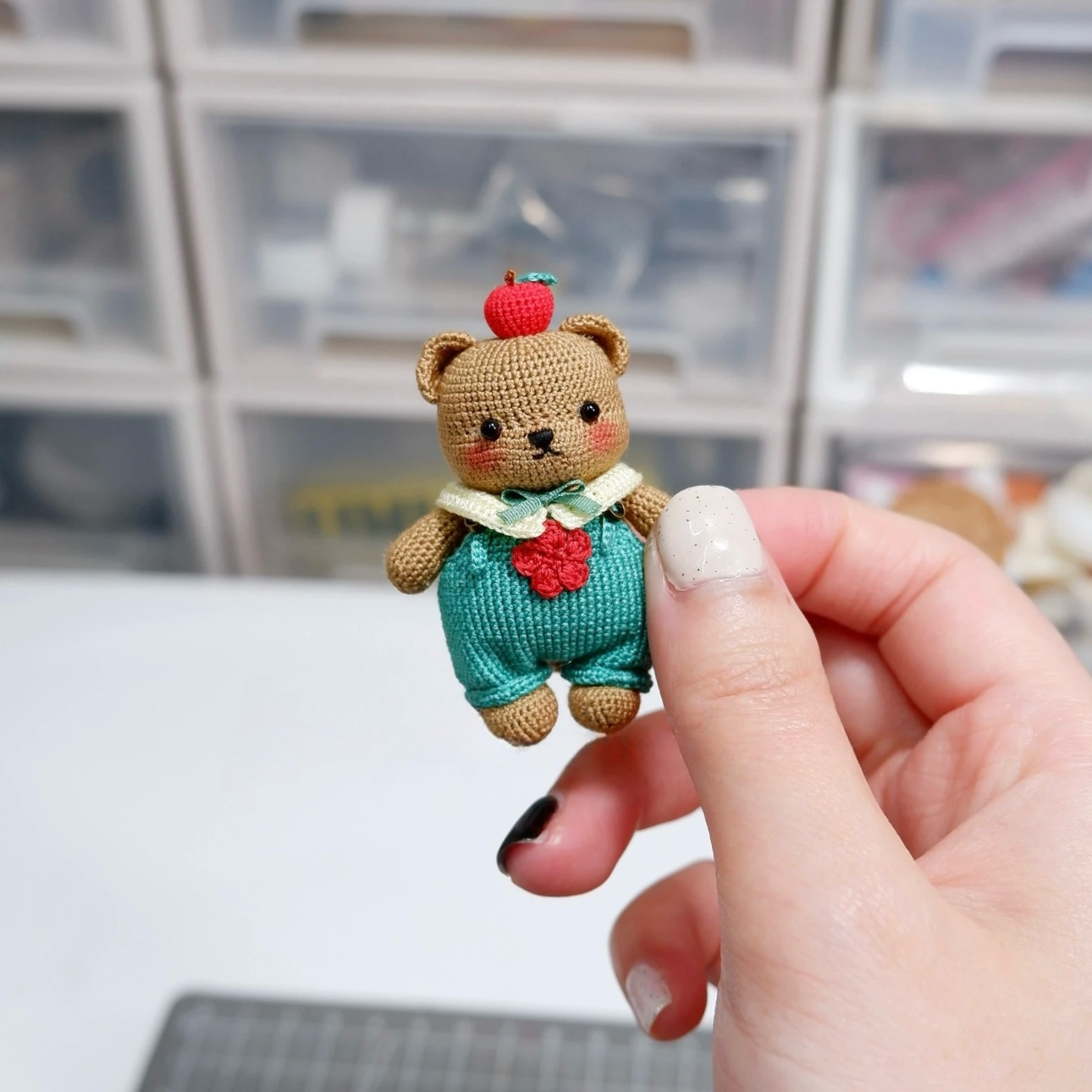 Hand holding a small teddy bear figurine with a red hat and green outfit against a blurred indoor background.