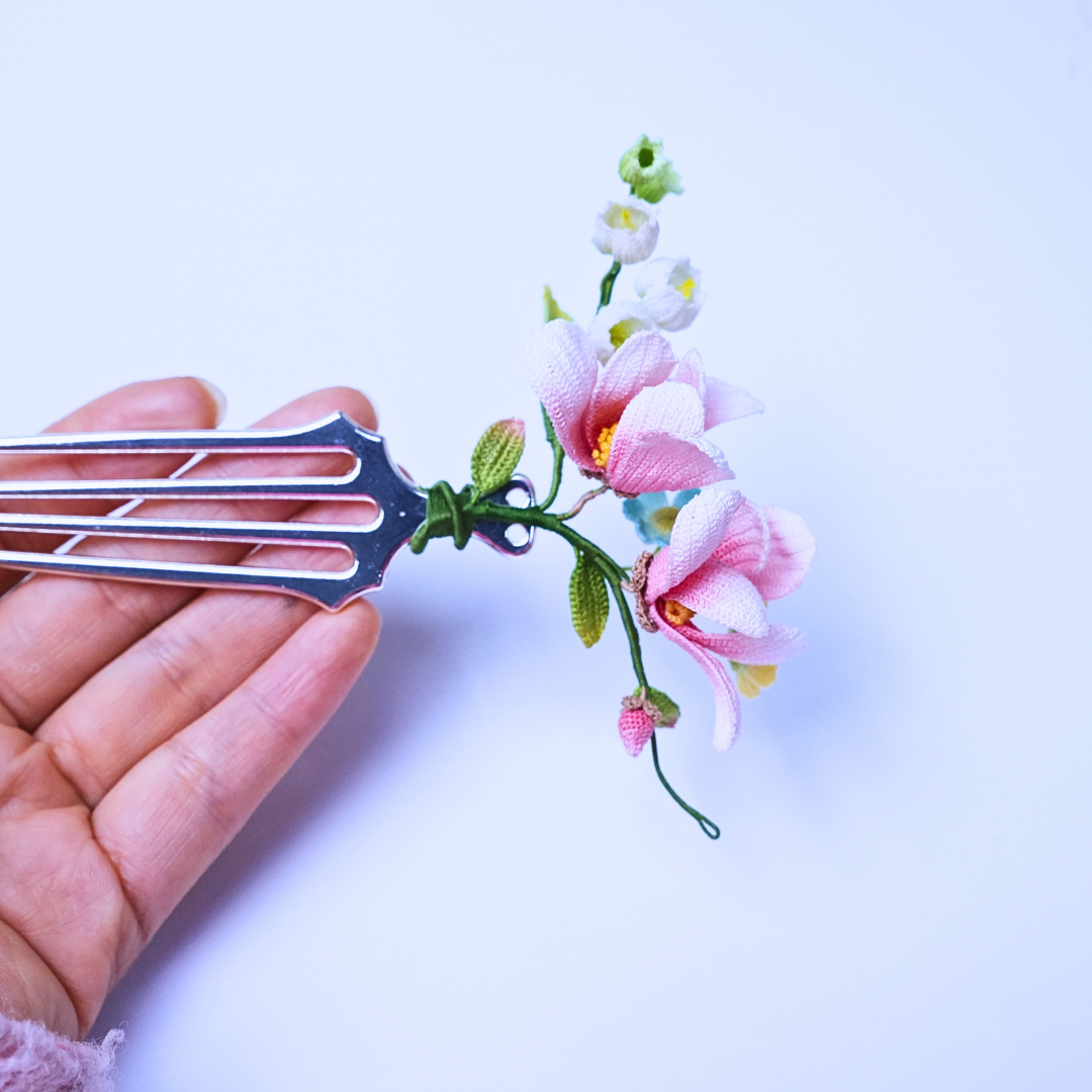 Handheld view of a micro crochet magnolia hair comb with a metal comb base and pink floral design