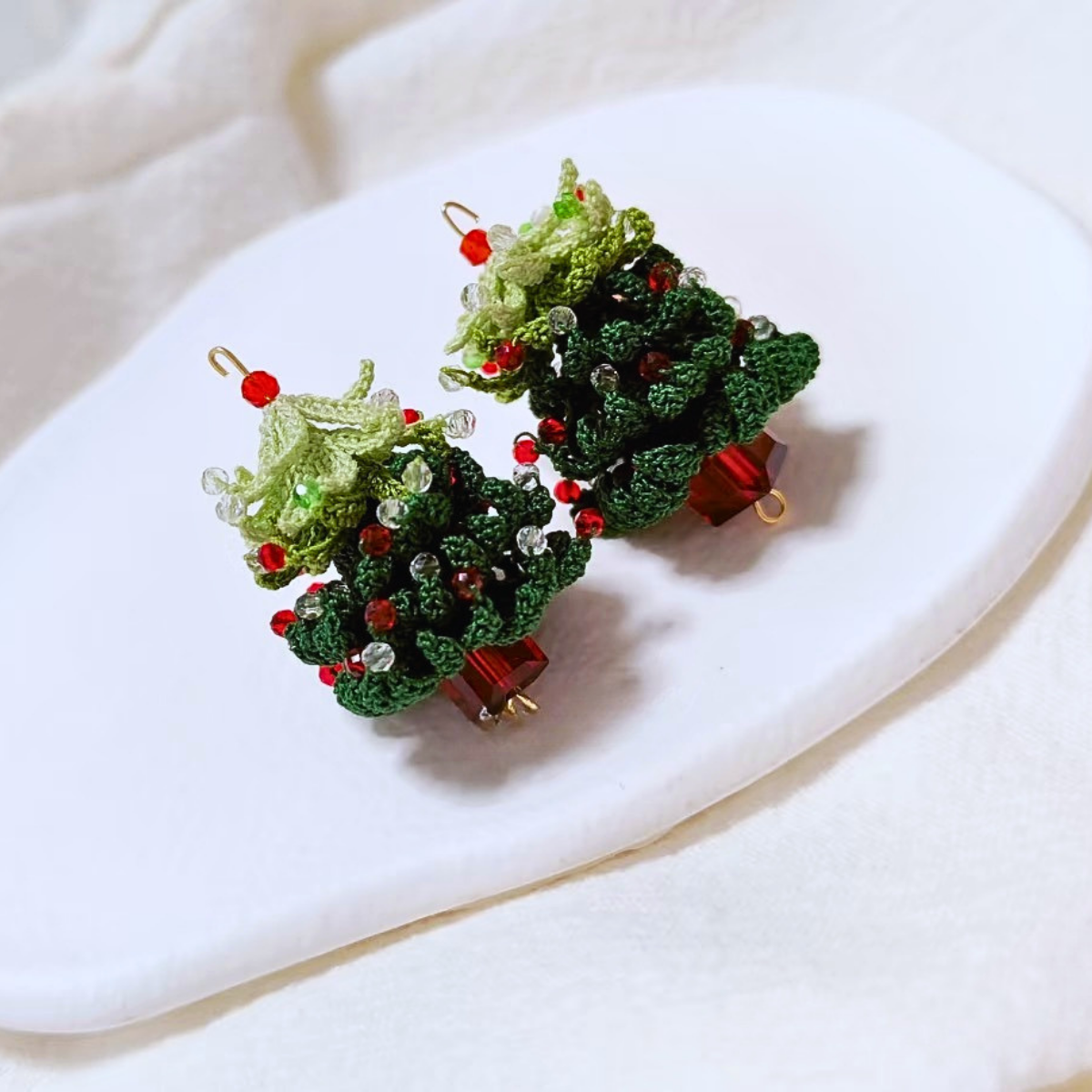 Pair of short-style handmade micro crochet Christmas tree earrings displayed on a white ceramic tray, showing layered green branches and red and clear bead details.