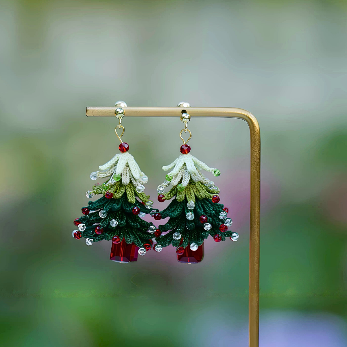Short-style handmade micro crochet Christmas tree earrings displayed on a gold-colored stand, with layered green branches and red and clear bead accents.